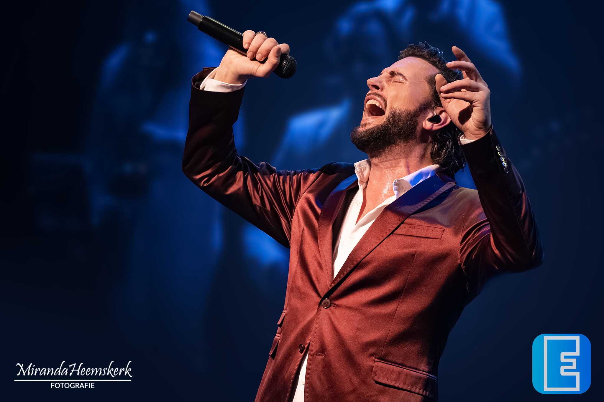 Een zanger treedt op onder blauwe podiumverlichting en houdt een microfoon omhoog. Hij draagt een rood pak met een wit overhemd en maakt een expressief handgebaar.