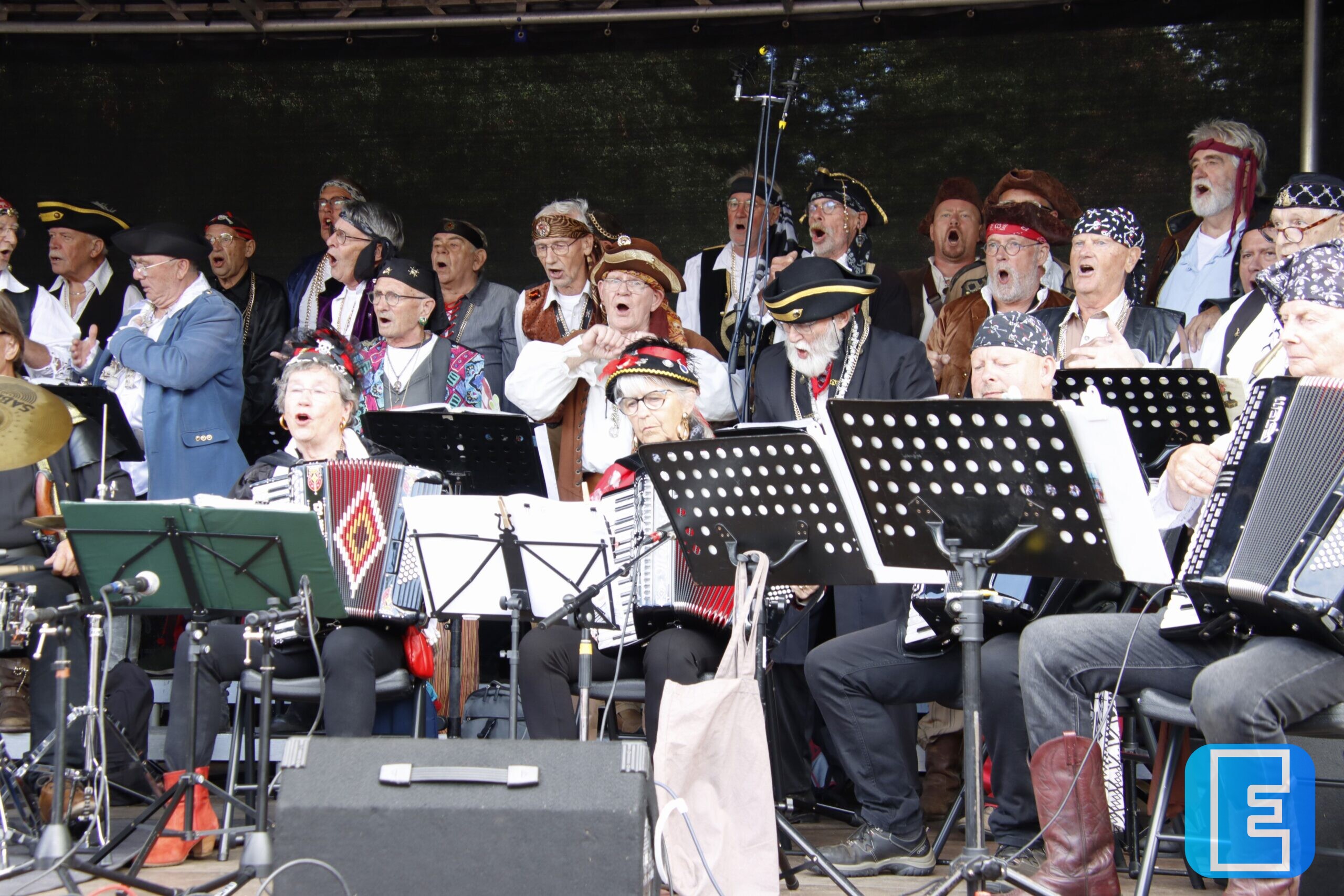 Een groot muzikaal ensemble treedt op onder een overkapping. Op de voorgrond zijn zangers en instrumentalisten te zien met accordeons, slagwerk, microfoons, muziekstandaards en bladmuziek.