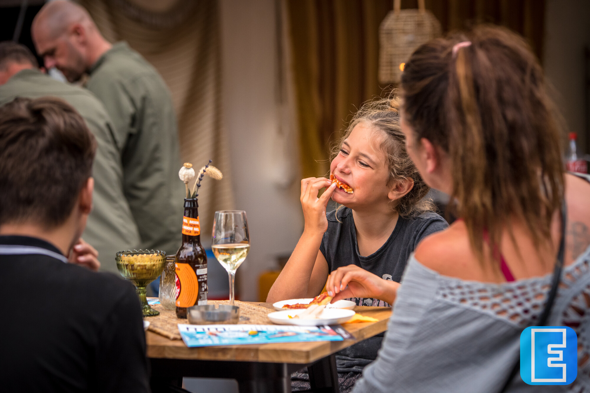 Gezelschap aan een gedekte tafel in een horecagelegenheid. Een vrouw eet terwijl op tafel borden, glazen, bestek en flessen staan.