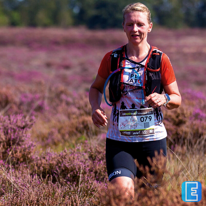 Een persoon in hardloopkleding rent door een uitgestrekt veld met paarse heide. De loper draagt een sportief drinkvest en een startnummer op de borst.