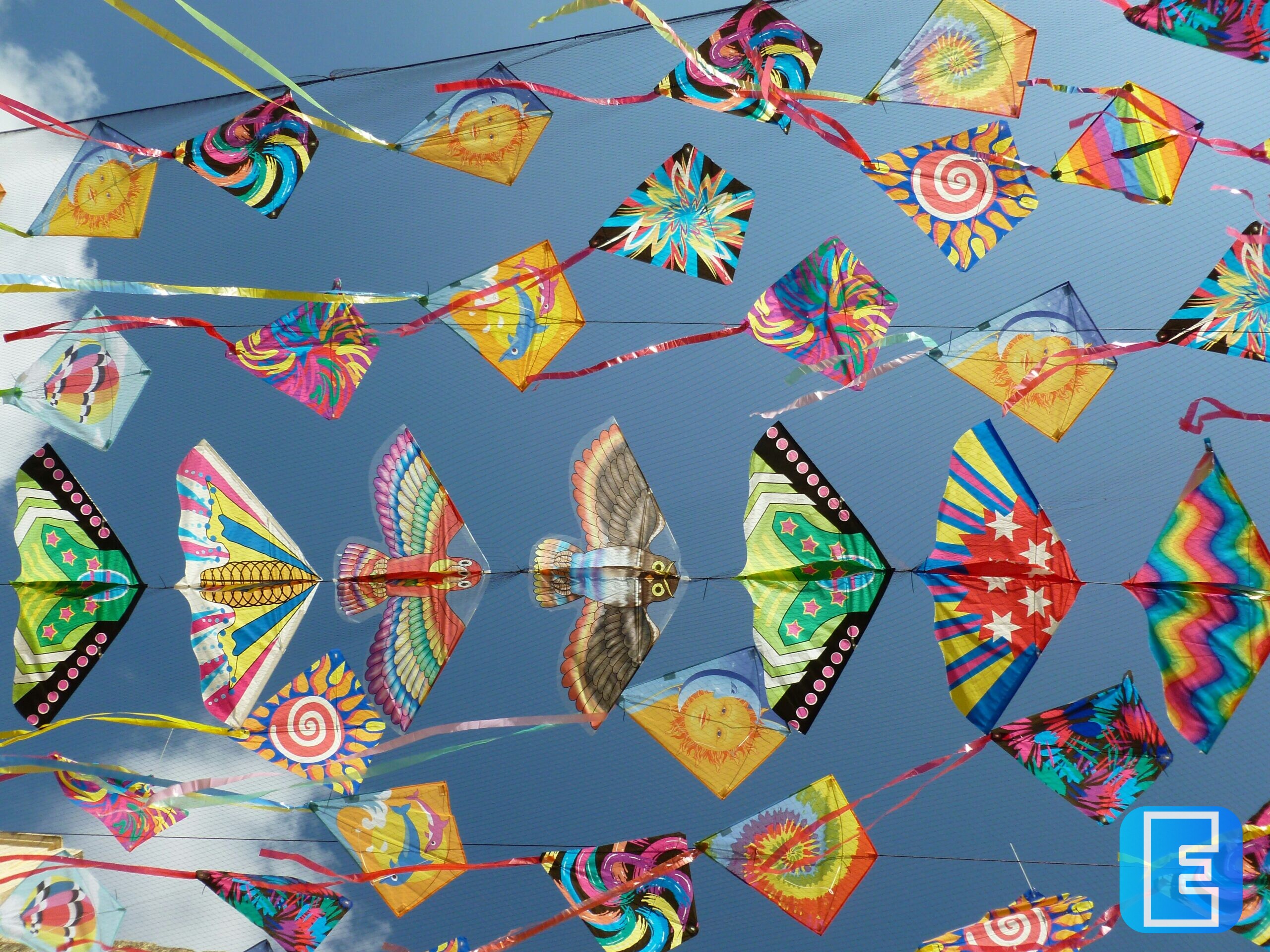 Tegen een blauwe lucht hangen veel kleurrijke vliegers en decoratieve waaiers, verbonden met slingers en draden. De vliegers hebben uiteenlopende patronen, waaronder spiralen, strepen en bloemachtige vormen.