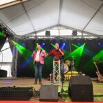 Twee artiesten staan op een podium onder een festivaltent en treden op voor een microfoon. Op het podium zijn gekleurde lichtbundels, muziekinstrumenten, luidsprekers en geluidsapparatuur zichtbaar.