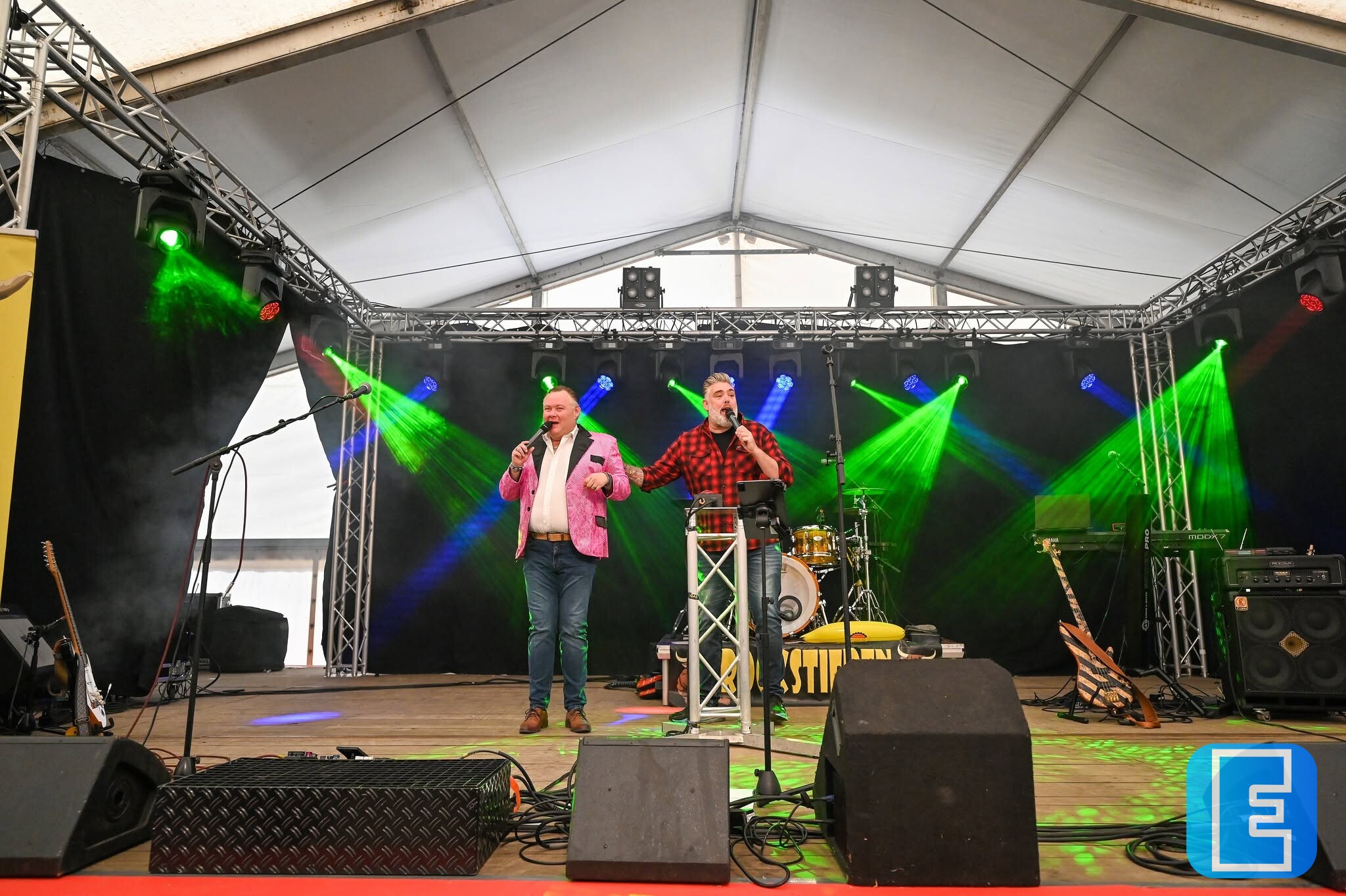 Twee artiesten staan op een podium onder een festivaltent en treden op voor een microfoon. Op het podium zijn gekleurde lichtbundels, muziekinstrumenten, luidsprekers en geluidsapparatuur zichtbaar.