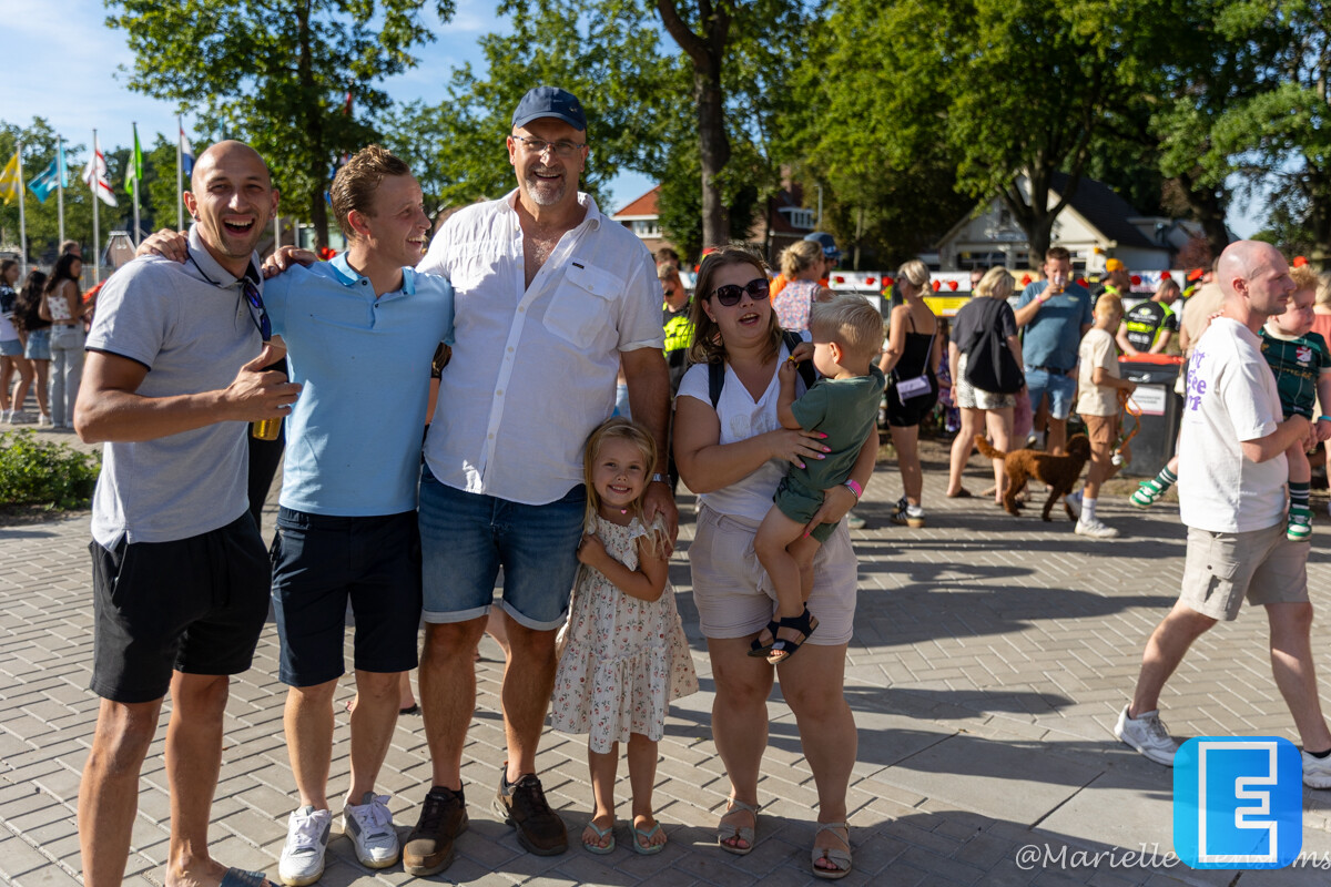 Een groep volwassenen en kinderen poseert lachend op een betegeld plein tijdens een druk bezocht buitenevenement. Op de achtergrond zijn vlaggen, bomen, kraampjes en andere bezoekers zichtbaar.