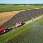 Luchtfoto van twee rode brandweerwagens op een smalle weg tussen uitgestrekte landbouwvelden. Enkele personen staan naast de voertuigen; er is geen brand zichtbaar.