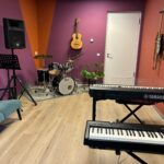 Interieur van een muzieklokaal met een drumstel, twee elektronische keyboards, een gitaar aan de muur, een luidspreker en een muziekstandaard. In de ruimte staan ook een bank en enkele planten.